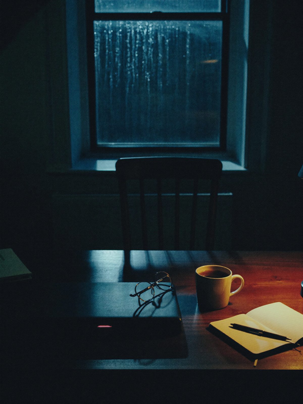 A kitchen table late at night: a closed laptop, a mug of tea, reading glasses and a notebook, rain on the window behind.